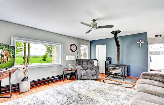 living room with baseboard heating, a wood stove, ceiling fan, and wood-type flooring