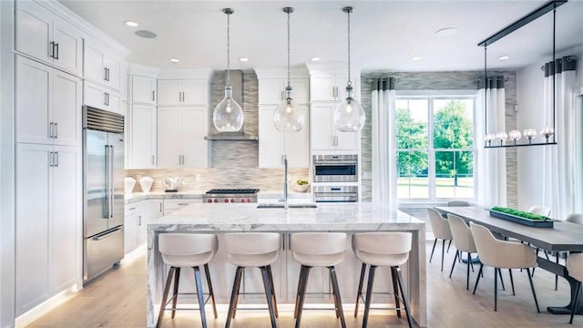 kitchen with stainless steel appliances, a kitchen island with sink, sink, wall chimney range hood, and light hardwood / wood-style flooring