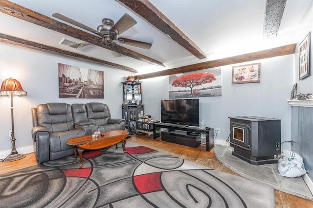 living room with beamed ceiling, wood-type flooring, a wood stove, and ceiling fan