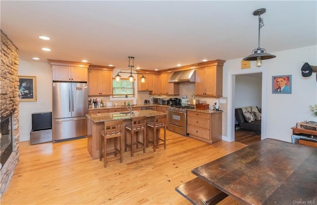 kitchen featuring appliances with stainless steel finishes, a kitchen breakfast bar, wall chimney exhaust hood, light hardwood / wood-style flooring, and a stone fireplace