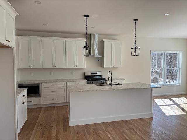 kitchen with stainless steel gas range oven, wall chimney range hood, a kitchen island with sink, and pendant lighting