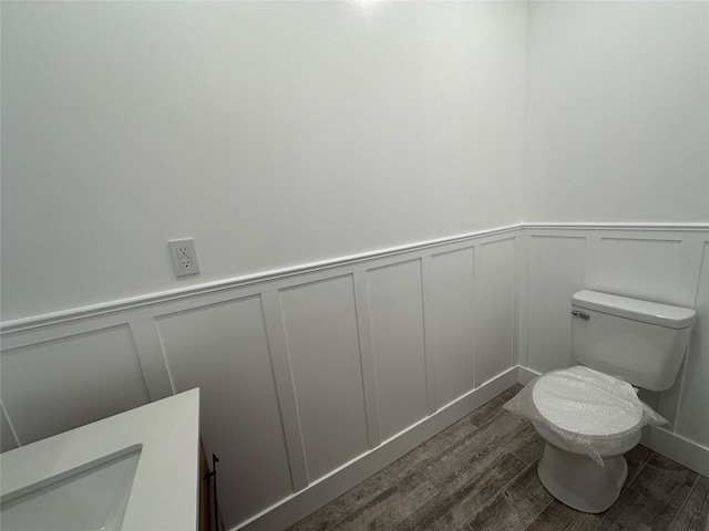 half bath with wood finished floors, a decorative wall, toilet, and wainscoting