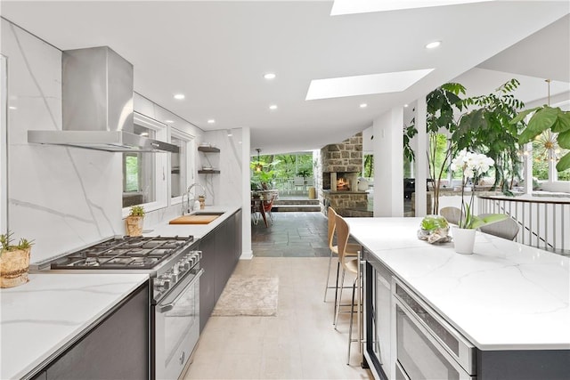 kitchen featuring a skylight, sink, wall chimney exhaust hood, light stone counters, and high end stove