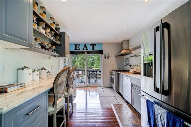 kitchen with decorative backsplash, appliances with stainless steel finishes, light stone counters, wall chimney exhaust hood, and hardwood / wood-style floors