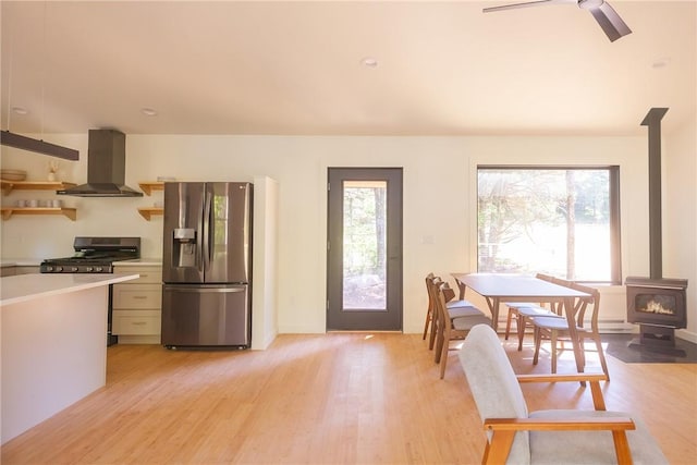 kitchen with wall chimney range hood, stainless steel fridge with ice dispenser, light hardwood / wood-style floors, a wood stove, and range with gas stovetop