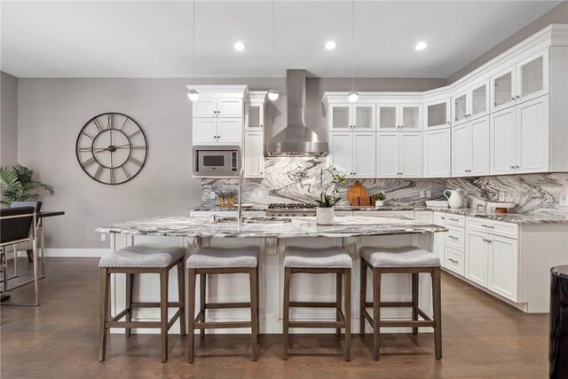 kitchen with hanging light fixtures, an island with sink, stainless steel microwave, and wall chimney range hood