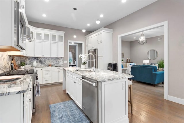 kitchen with light stone countertops, stainless steel appliances, dark wood-type flooring, white cabinetry, and an island with sink