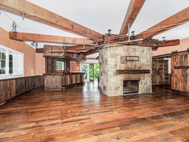 unfurnished living room featuring beam ceiling, a stone fireplace, a barn door, hardwood / wood-style floors, and wooden walls