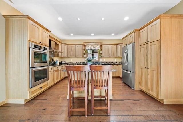 kitchen featuring dark wood-style flooring, wall chimney range hood, light brown cabinetry, and appliances with stainless steel finishes