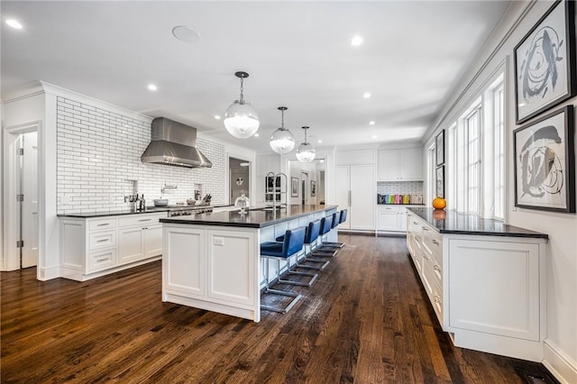 kitchen with wall chimney exhaust hood, dark wood-type flooring, hanging light fixtures, a center island with sink, and white cabinets