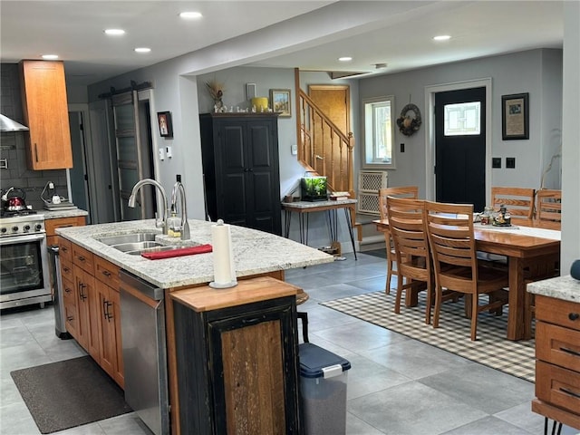 kitchen with sink, a barn door, an island with sink, appliances with stainless steel finishes, and light stone counters