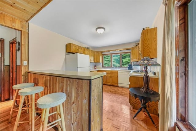 kitchen with kitchen peninsula, a kitchen bar, white appliances, light parquet flooring, and wood walls