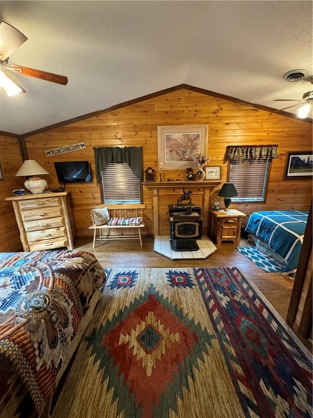 bedroom with wood walls, lofted ceiling, a wood stove, ceiling fan, and a textured ceiling