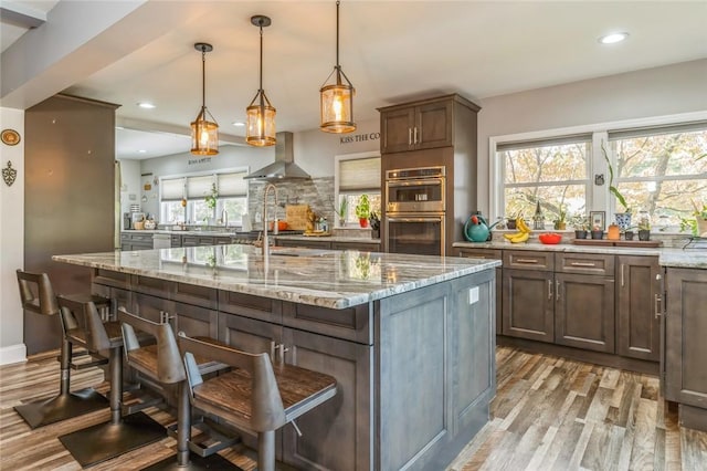 bar featuring light stone counters, wall chimney exhaust hood, stainless steel double oven, decorative light fixtures, and light hardwood / wood-style floors