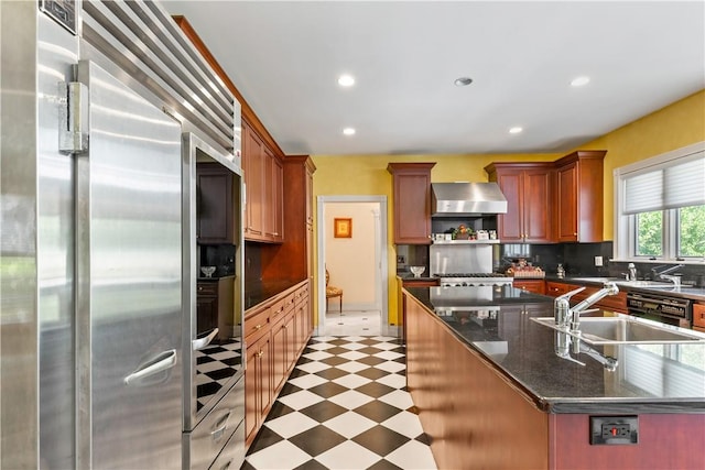 kitchen with built in fridge, a center island with sink, wall chimney range hood, sink, and tasteful backsplash