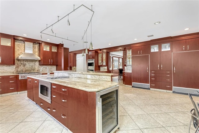 kitchen with a large island with sink, wall chimney range hood, appliances with stainless steel finishes, decorative light fixtures, and beverage cooler