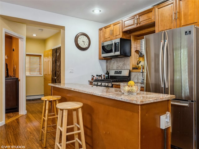 kitchen with appliances with stainless steel finishes, a baseboard radiator, dark wood finished floors, backsplash, and light stone counters