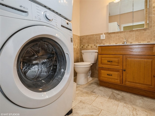 bathroom featuring stacked washing machine and dryer, tile walls, toilet, vanity, and stone tile flooring