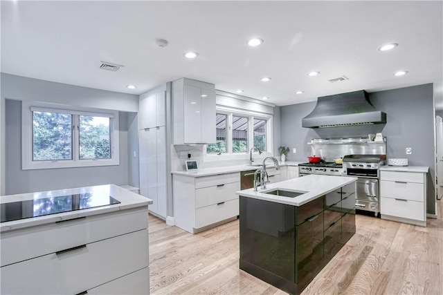 kitchen featuring white cabinetry, stainless steel range, wall chimney range hood, an island with sink, and black electric cooktop