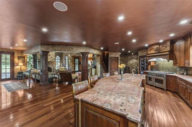 kitchen featuring custom exhaust hood, a large island, light stone counters, wood-type flooring, and decorative columns