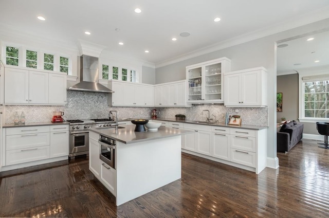 kitchen featuring wall chimney exhaust hood, sink, white cabinetry, appliances with stainless steel finishes, and a kitchen island with sink