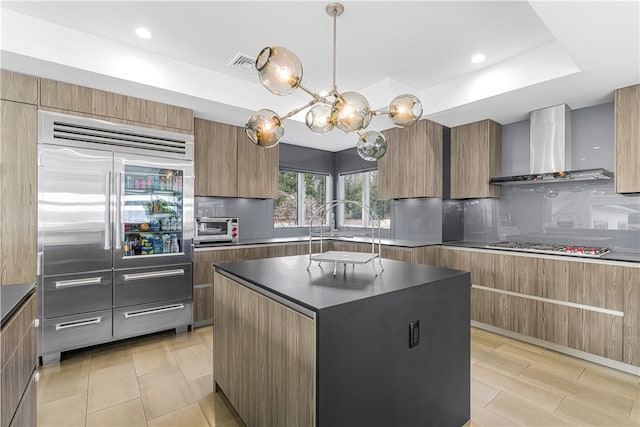 kitchen with wall chimney exhaust hood, hanging light fixtures, a raised ceiling, a kitchen island, and appliances with stainless steel finishes