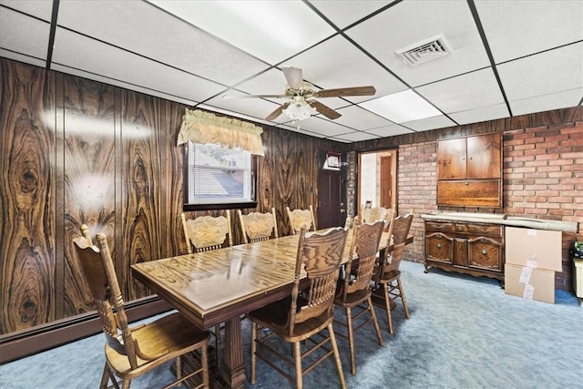 carpeted dining space featuring baseboard heating, a drop ceiling, wooden walls, and ceiling fan