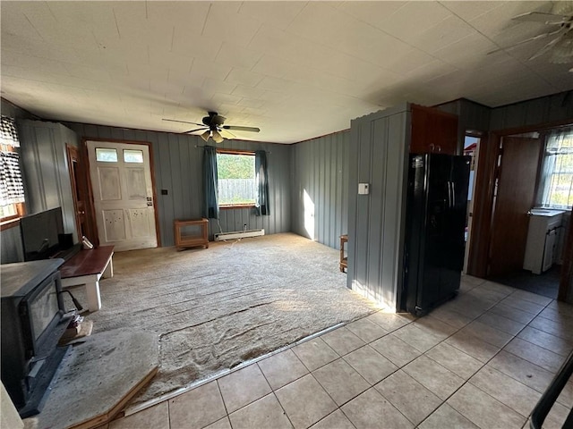 unfurnished living room with baseboard heating, a wood stove, ceiling fan, and light tile patterned flooring