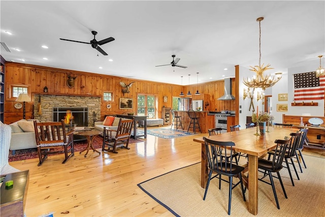 dining room featuring ceiling fan with notable chandelier, a stone fireplace, wooden walls, light hardwood / wood-style flooring, and ornamental molding