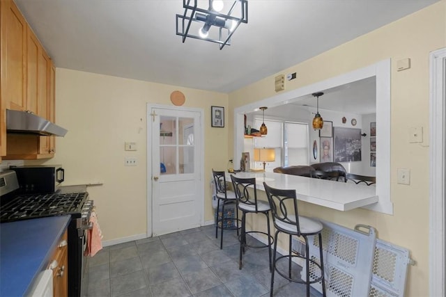 kitchen featuring light brown cabinetry, stainless steel gas range, dark tile patterned flooring, hanging light fixtures, and a breakfast bar area