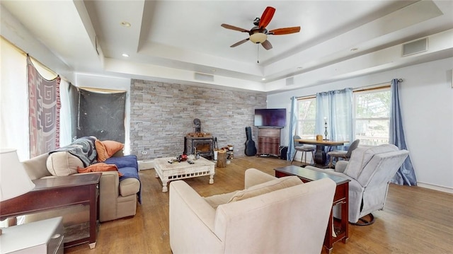 living room with ceiling fan, a raised ceiling, a wood stove, and light hardwood / wood-style flooring