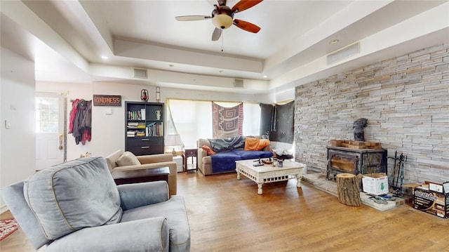 living room featuring wood-type flooring, a tray ceiling, a wood stove, and ceiling fan