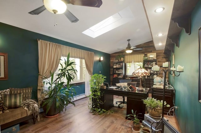 home office featuring ceiling fan, baseboard heating, wood walls, vaulted ceiling with skylight, and hardwood / wood-style flooring