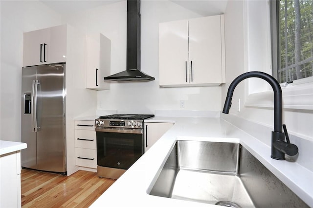 kitchen featuring wall chimney range hood, sink, appliances with stainless steel finishes, light hardwood / wood-style floors, and white cabinetry