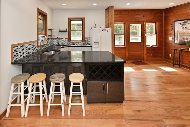 kitchen with kitchen peninsula, dark stone counters, wooden walls, white refrigerator with ice dispenser, and light hardwood / wood-style flooring