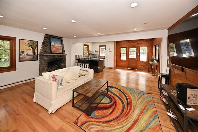 living room featuring a stone fireplace, light wood-type flooring, baseboard heating, and wooden walls