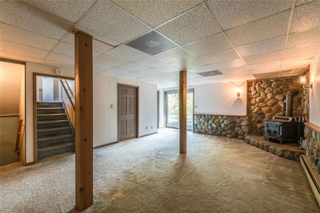 basement with baseboard heating, a wood stove, a drop ceiling, and carpet