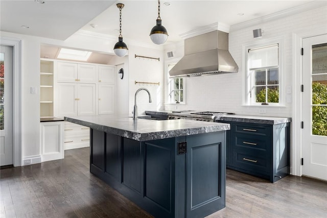 kitchen with sink, wall chimney range hood, decorative light fixtures, dark hardwood / wood-style floors, and an island with sink