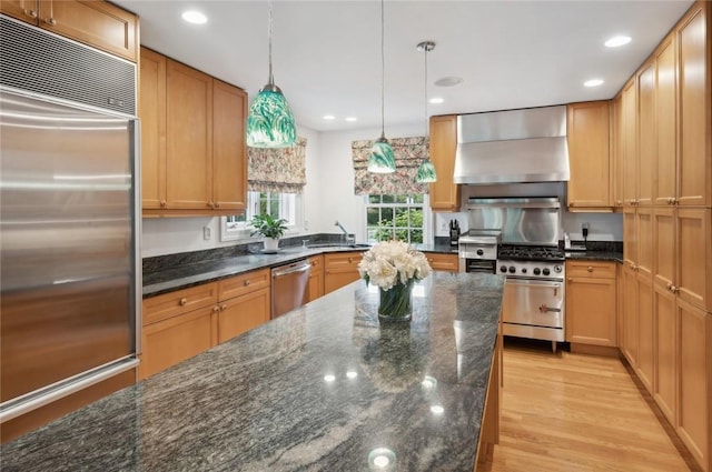 kitchen with wall chimney range hood, light hardwood / wood-style flooring, dark stone countertops, pendant lighting, and high quality appliances