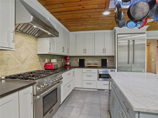 kitchen with wall chimney range hood, premium appliances, dark stone counters, decorative backsplash, and white cabinets