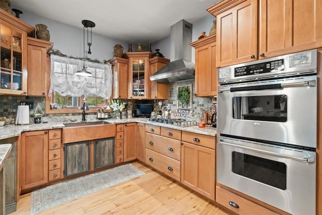 kitchen with stainless steel appliances, wall chimney range hood, light stone counters, light hardwood / wood-style floors, and decorative backsplash