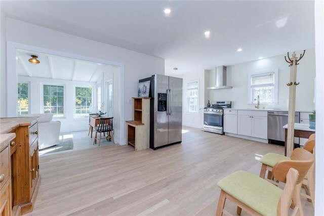 kitchen featuring appliances with stainless steel finishes, light wood-type flooring, wall chimney range hood, lofted ceiling with beams, and white cabinetry