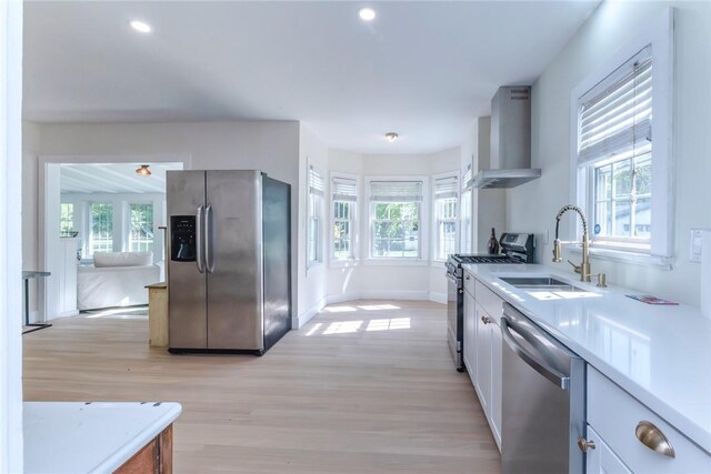 kitchen with light wood-type flooring, stainless steel appliances, sink, wall chimney range hood, and white cabinets