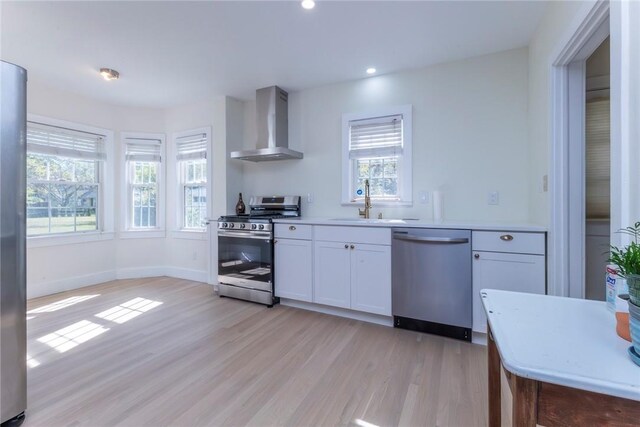 kitchen featuring a healthy amount of sunlight, stainless steel appliances, wall chimney range hood, and light hardwood / wood-style floors