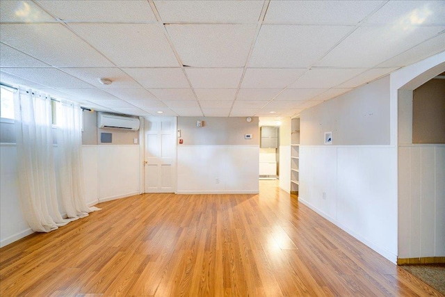 basement with a drop ceiling, a wall mounted air conditioner, and light wood-type flooring