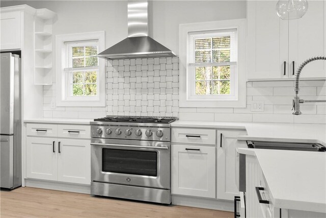 kitchen with light wood-type flooring, stainless steel appliances, wall chimney range hood, pendant lighting, and white cabinetry