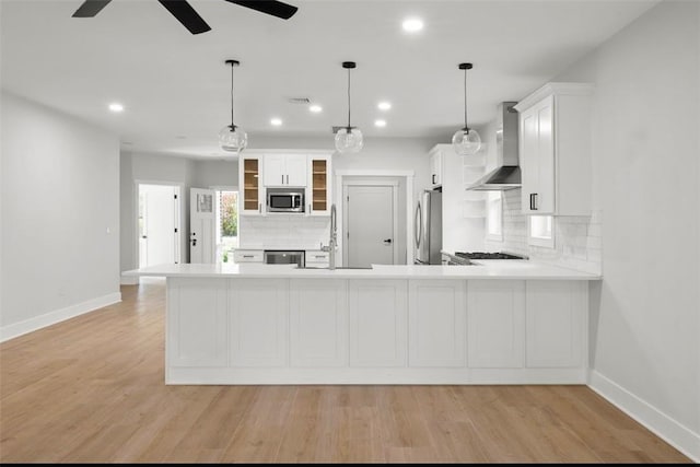 kitchen featuring stainless steel appliances, wall chimney range hood, kitchen peninsula, pendant lighting, and light hardwood / wood-style floors