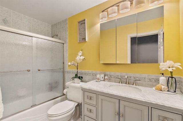 full bath with tile walls, bath / shower combo with glass door, toilet, wainscoting, and vanity