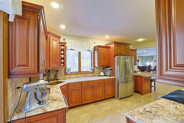 kitchen with light stone counters, a sink, backsplash, freestanding refrigerator, and brown cabinetry