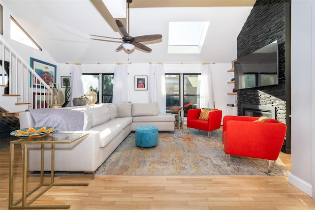 living room with a stone fireplace, ceiling fan, light hardwood / wood-style flooring, and vaulted ceiling with skylight